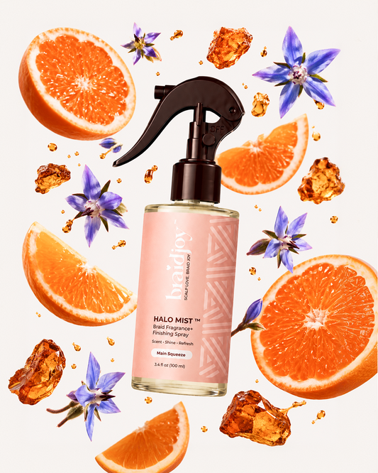 Braidjoy Halo Mist braid fragrance and finishing spray in Main Squeeze, surrounded by orange slices, purple flowers, and amber droplets, designed to refresh braids, neutralize odor, and add a lightweight salon-fresh shine.