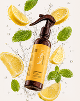 Amber spray bottle of braidjoy Clean Sweep dandruff and buildup eraser surrounded by splashing water, lemon slices, and mint leaves, highlighting a refreshing pre-shampoo scalp treatment for buildup, dandruff, and cleaner wash days.