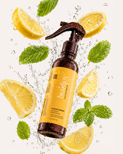 Amber spray bottle of braidjoy Clean Sweep dandruff and buildup eraser surrounded by splashing water, lemon slices, and mint leaves, highlighting a refreshing pre-shampoo scalp treatment for buildup, dandruff, and cleaner wash days.